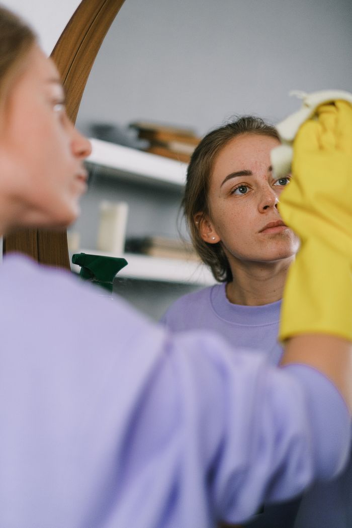 Femme coupée au cadrage, en sweatshirt violet et gants en latex, nettoyant un miroir dans un appartement lumineux en journée